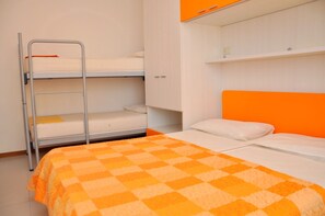 En køjeseng med et ternet orange og hvidt tæppe, en metalstige og en hvid væg med en hylde over.