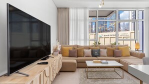 Un salon moderne doté d’un grand téléviseur à écran plat, d’un canapé d’angle beige, d’une table basse et offrant une vue sur le paysage urbain grâce à de grandes fenêtres.