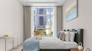 Une chambre moderne avec une grande fenêtre, un lit agrémenté de plusieurs coussins, une table de chevet avec une lampe et une petite table sur laquelle se trouve un bol de fruits.