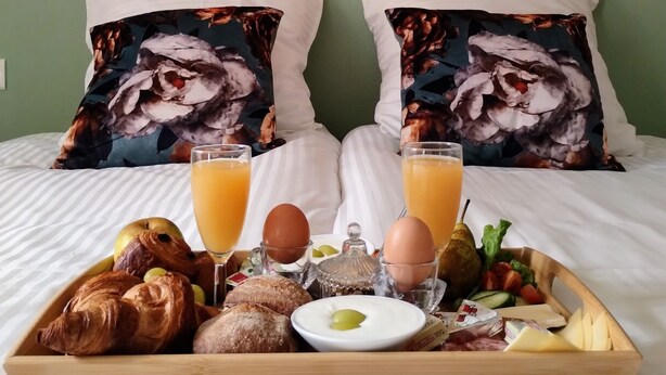 Un lit bien fait, avec deux oreillers, un plateau de petit-déjeuner comprenant des croissants, des œufs et du jus, et un tableau représentant un paysage fluvial au-dessus du lit.