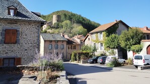Un bâtiment en pierre, doté d’un toit en pente et de volets en bois, entouré d’une rue pavée et bordé de voitures garées.