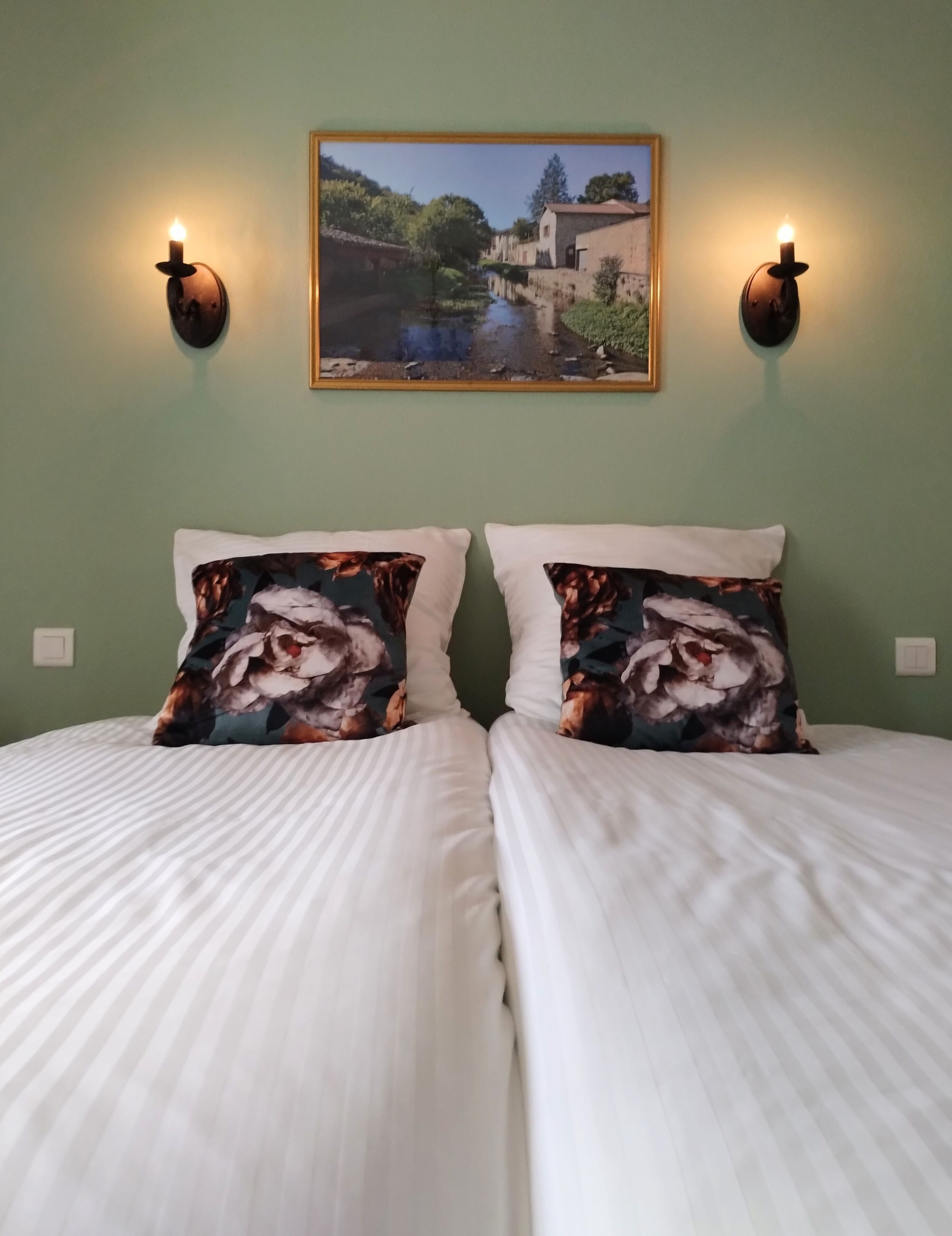 Un lit double avec du linge de lit blanc, deux coussins décoratifs à motifs floraux et, au-dessus, un tableau représentant un paysage encadré.