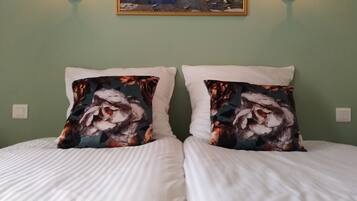Un lit double avec du linge de lit blanc, deux coussins décoratifs à motifs floraux et, au-dessus, un tableau représentant un paysage encadré.