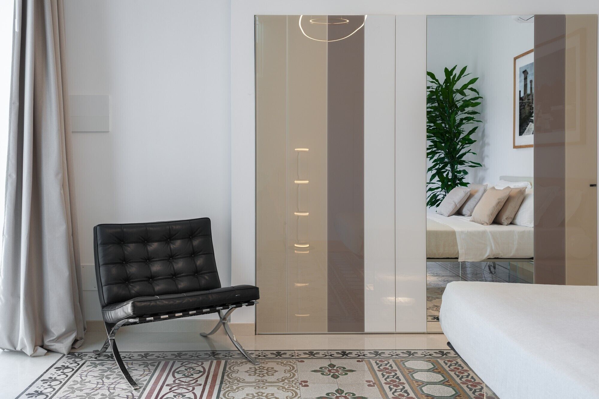 Un dormitorio moderno con una butaca de cuero negro, una cama con cubrecama blanco y un espejo grande con tiras de luz verticales.