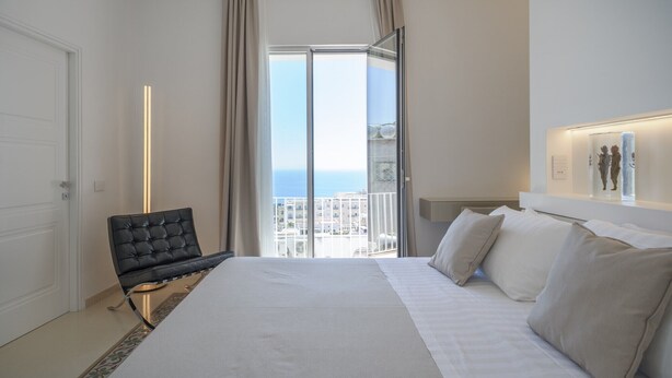Habitación de hotel con cama, una silla, un ventanal con vista a la ciudad y una lámpara fijada a la pared.
