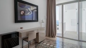 Una sala moderna con una obra de arte grande enmarcada, un escritorio de cristal, una silla, una alfombra estampada y un ventanal corredero que da a un balcón.