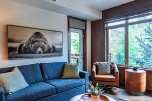 Ein modernes Wohnzimmer mit einem blauen Sofa, einem braunen Ledersessel und einem großen Fenster mit Blick auf Bäume.