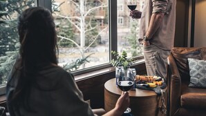 A person standing by a window holding a glass of wine, with a table in front of them containing a bottle, a glass, and a plant. - Three Bears Hydeaway at Stoneridge (Canmore)