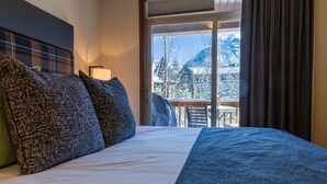 A hotel room with a large bed, a view of snow-covered mountains, and a balcony with a table and chairs. - Three Bears Hydeaway at Stoneridge (Canmore)