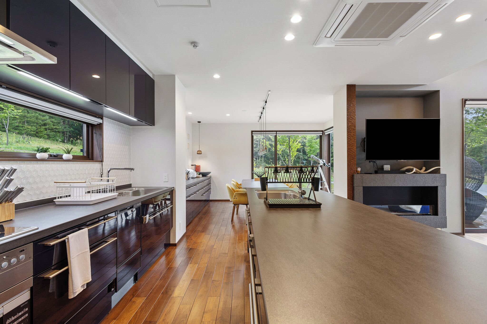 A modern kitchen with dark cabinetry, a wooden countertop, and a large window overlooking a garden.