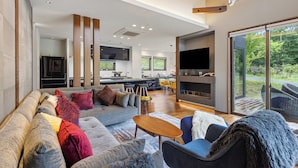 Una sala de estar moderna con sofá, sillones y televisión. Se observa una cocina al fondo, con un ventanal grande.