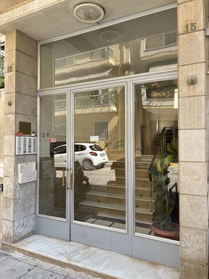 L’entrée d’un immeuble avec une porte en verre, une voiture blanche garée devant, et un escalier menant à un étage supérieur.