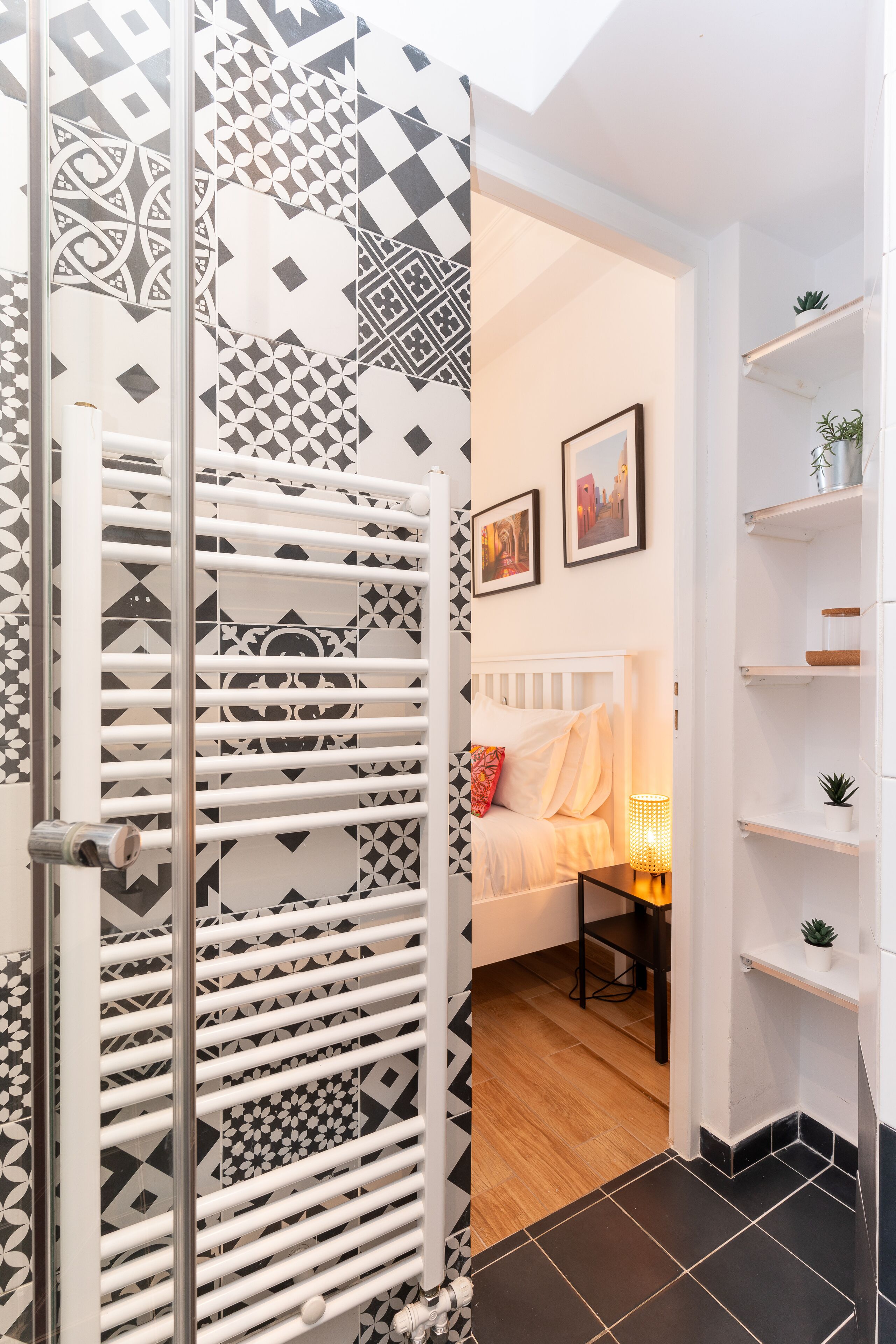 Um banheiro moderno com azulejos estampados, um radiador branco, uma cama com cabeceira branca e uma prateleira com plantas.