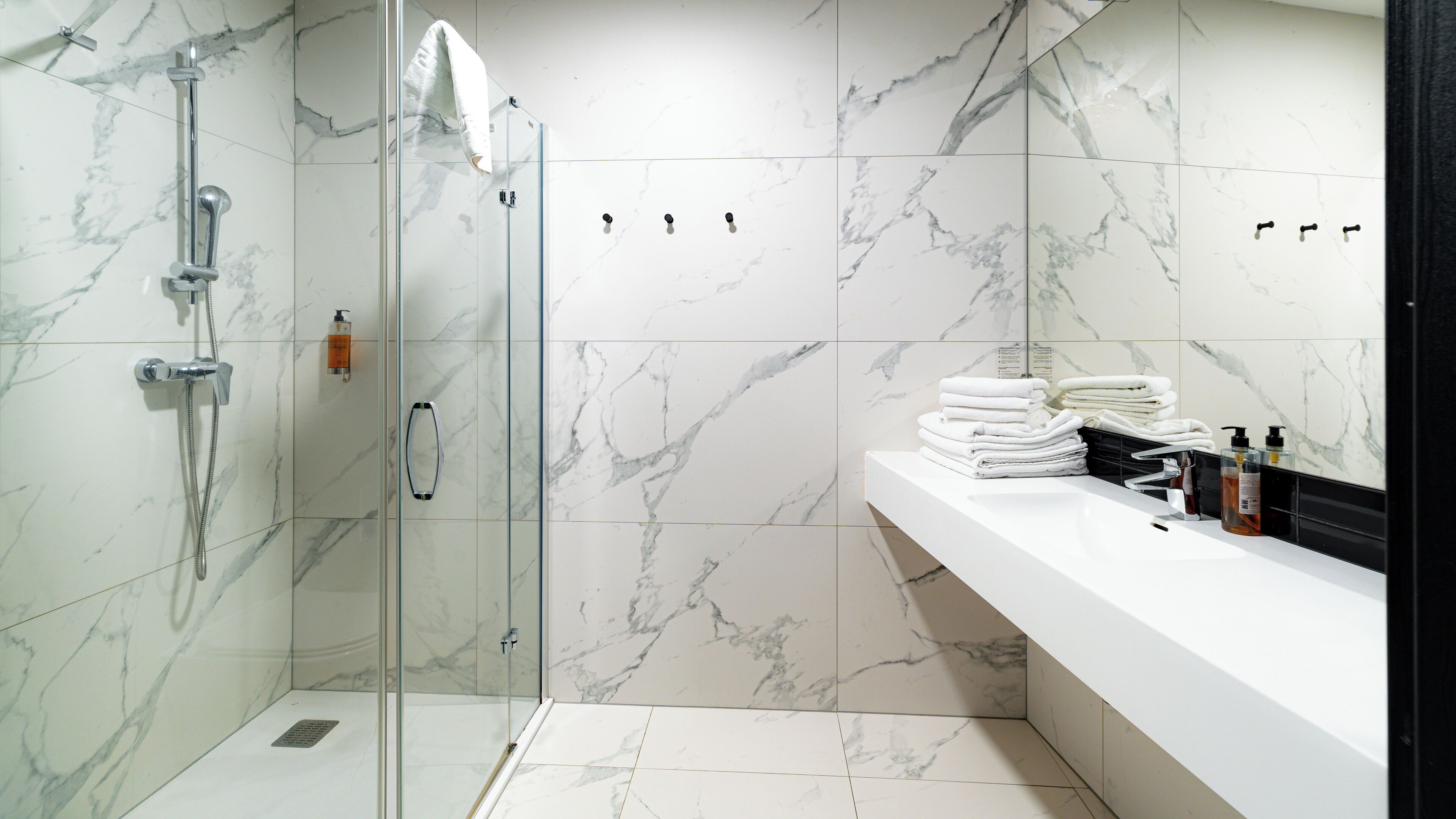 Un baño moderno con ducha envuelta en cristal, azulejos de mármol, un tocador blanco con artículos de aseo y un perchero.