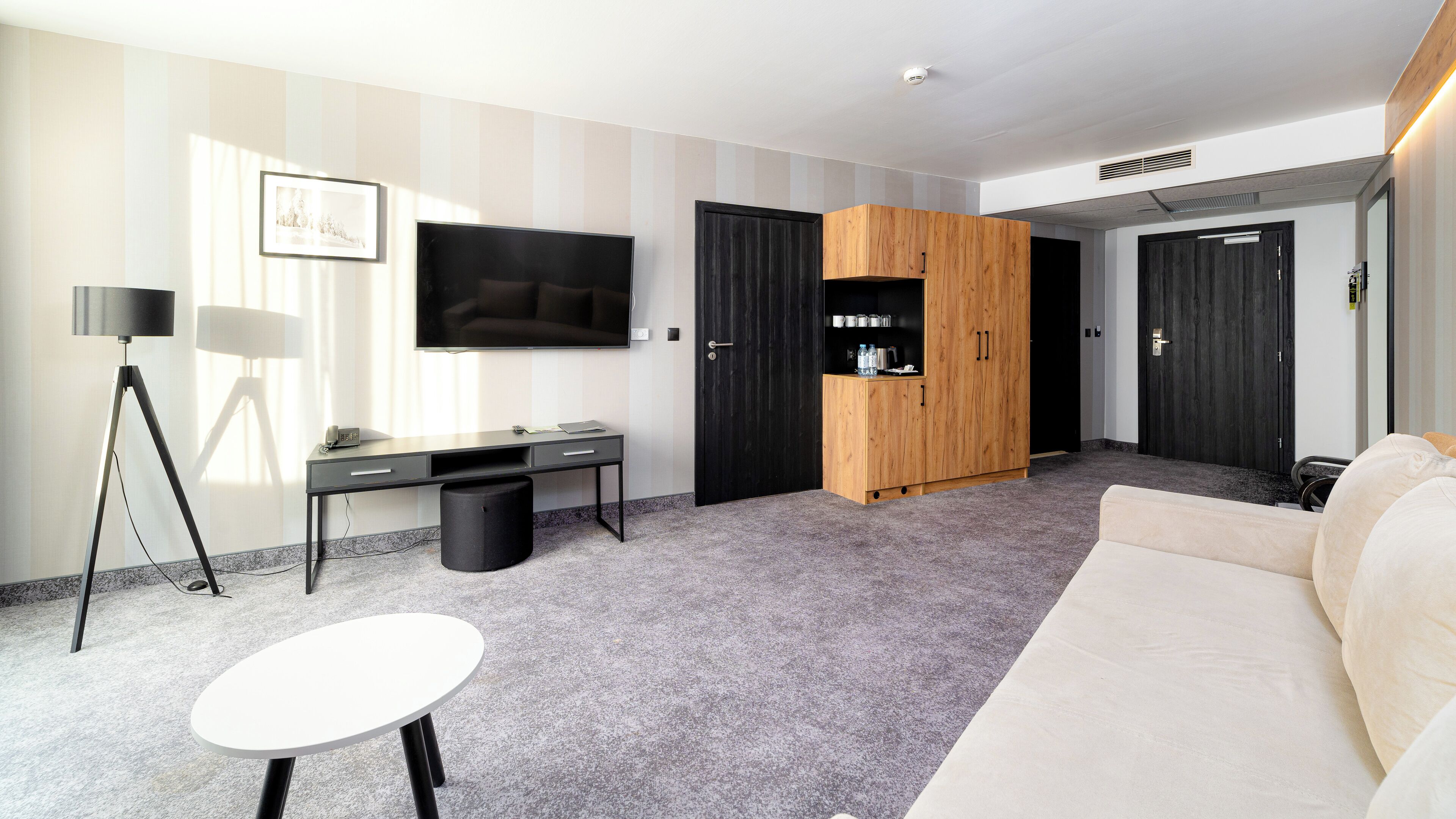 A modern hotel room with a flat-screen TV, a desk with a lamp, a sofa, and a wardrobe.