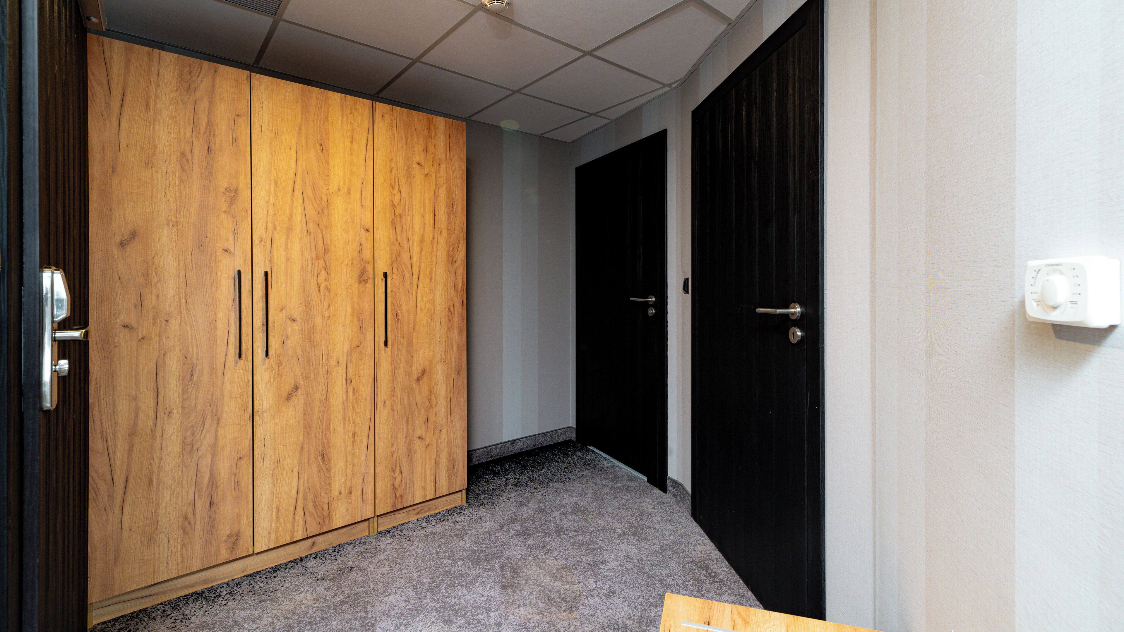 A hallway with a wooden wardrobe, a black door, and a thermostat on the wall.