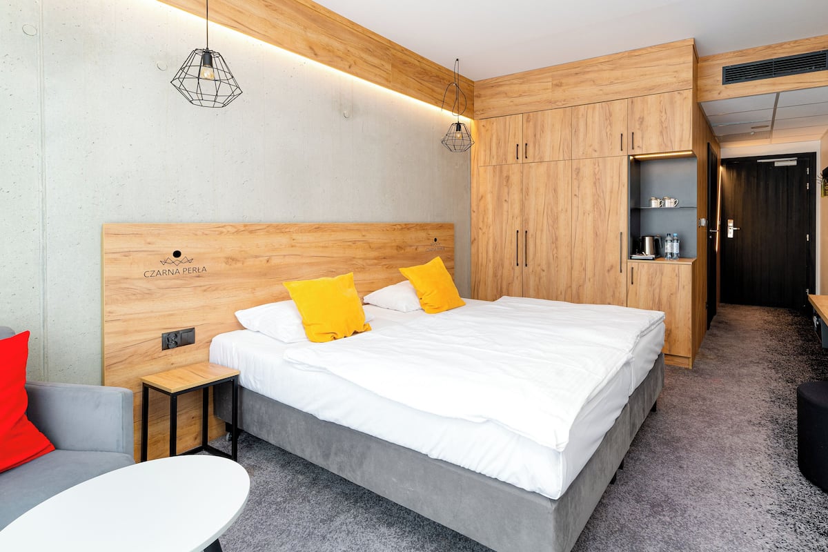 Una habitación de hotel moderna con cabecera de madera, cama con ropa blanca y almohadas amarillas, un escritorio con ordenador y un pequeño rincón de descanso con una almohada roja.