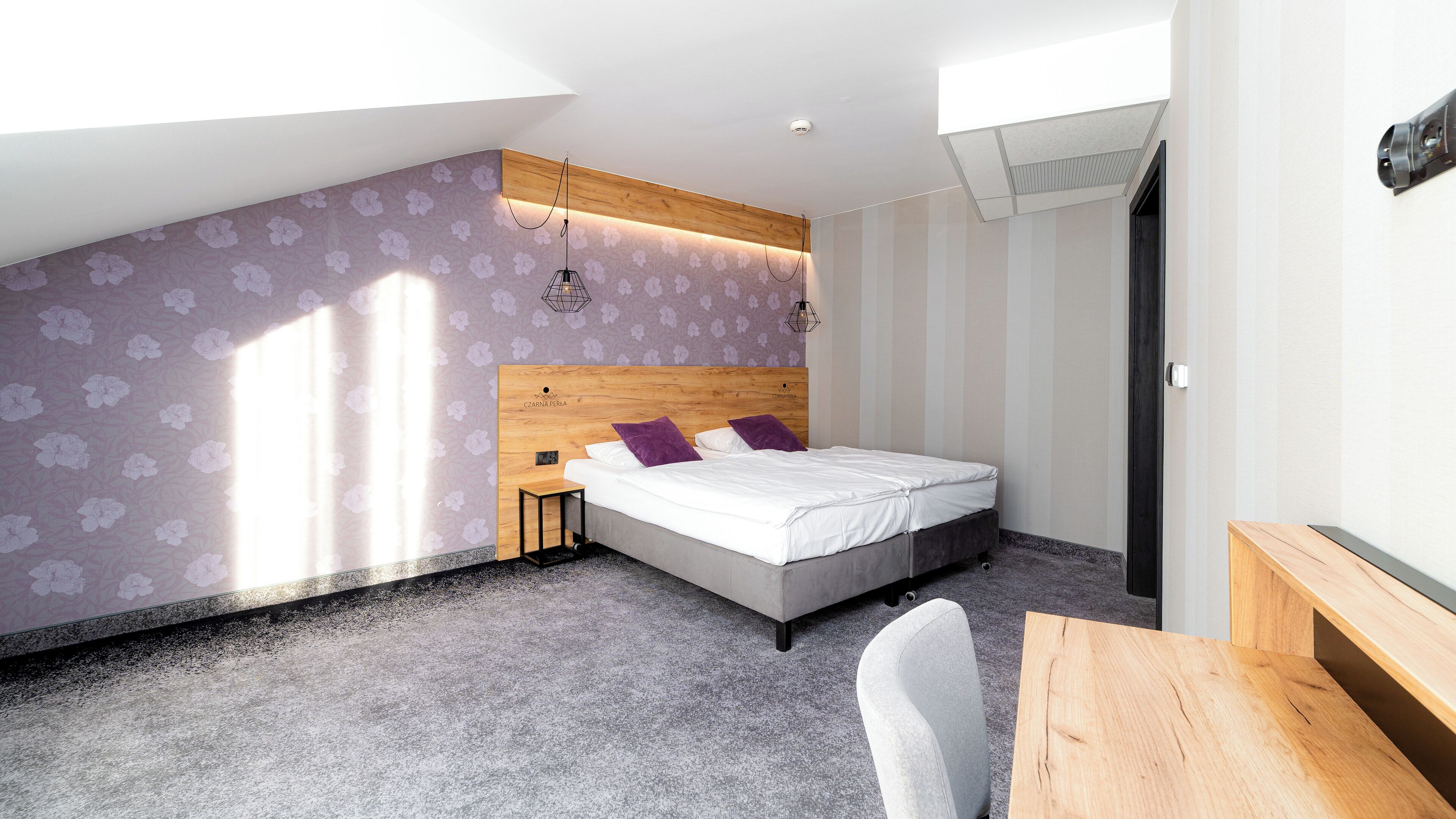 A modern bedroom with a sloped ceiling, a bed with white bedding and purple pillows, a wooden headboard, and a small bedside table.