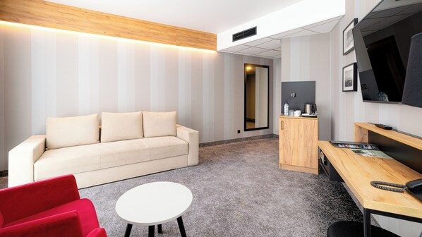 Une chambre d’hôtel moderne, équipée d’un canapé, d’une petite table ronde, d’un fauteuil rouge, d’un bureau en bois et d’une télévision.