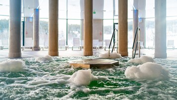 Zona de spa con una bañera de hidromasaje rodeada de espuma blanca, ventanas amplias y columnas.