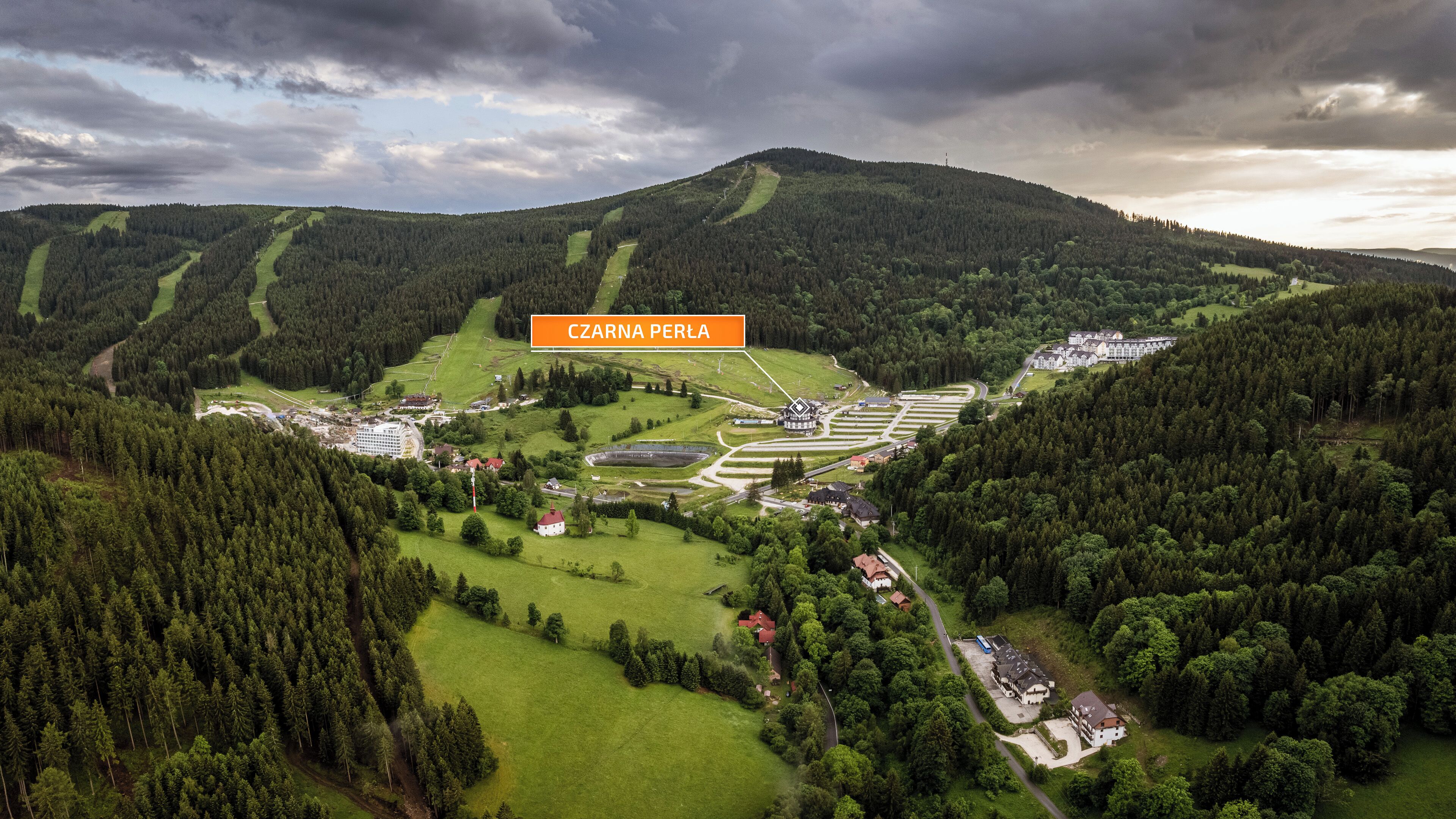 An aerial view of Zarna Perla, featuring a large hotel complex, surrounding forested areas, and a hilly landscape.