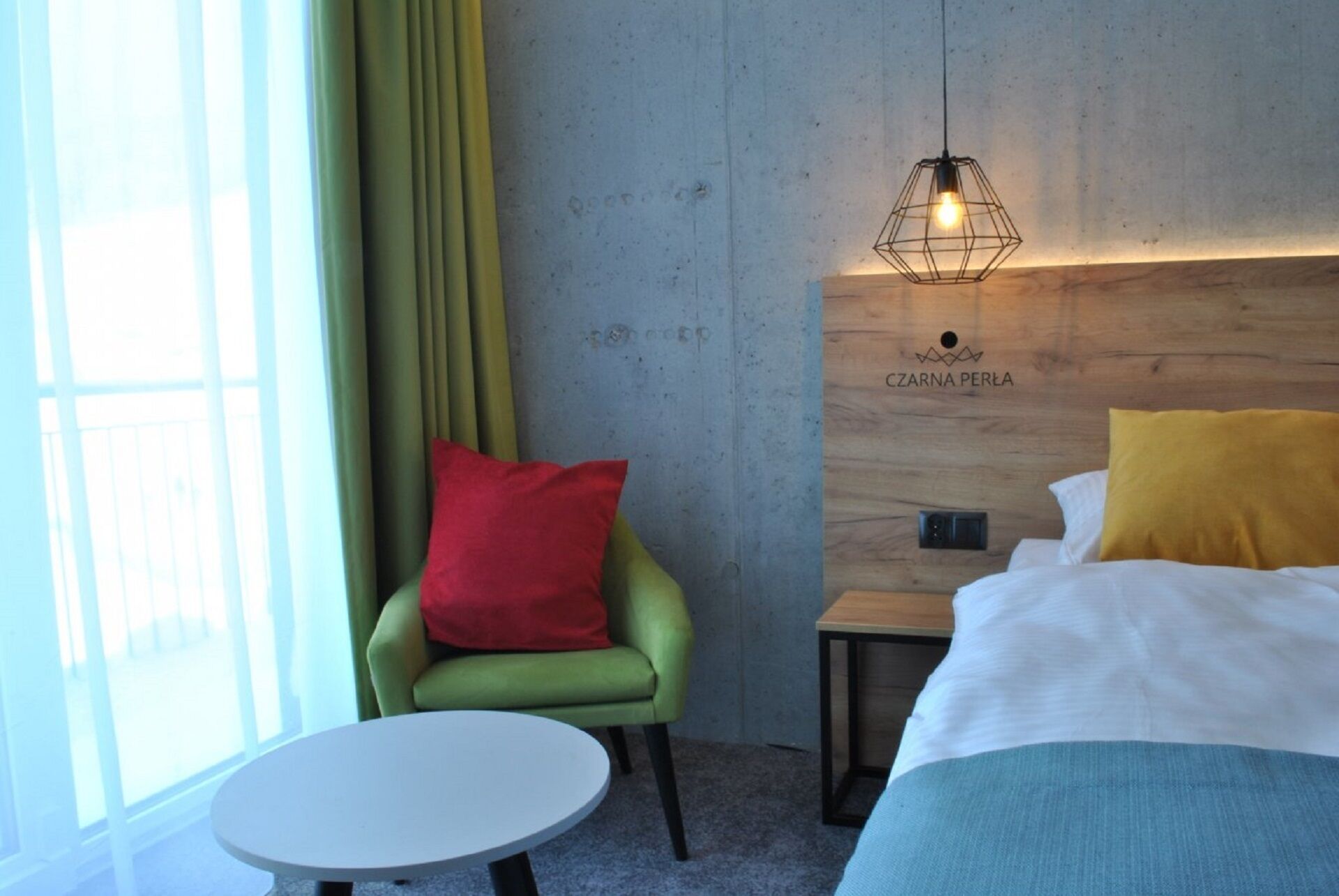 A hotel room with a bed, a green armchair, a small round table, and a wooden headboard with a light fixture.