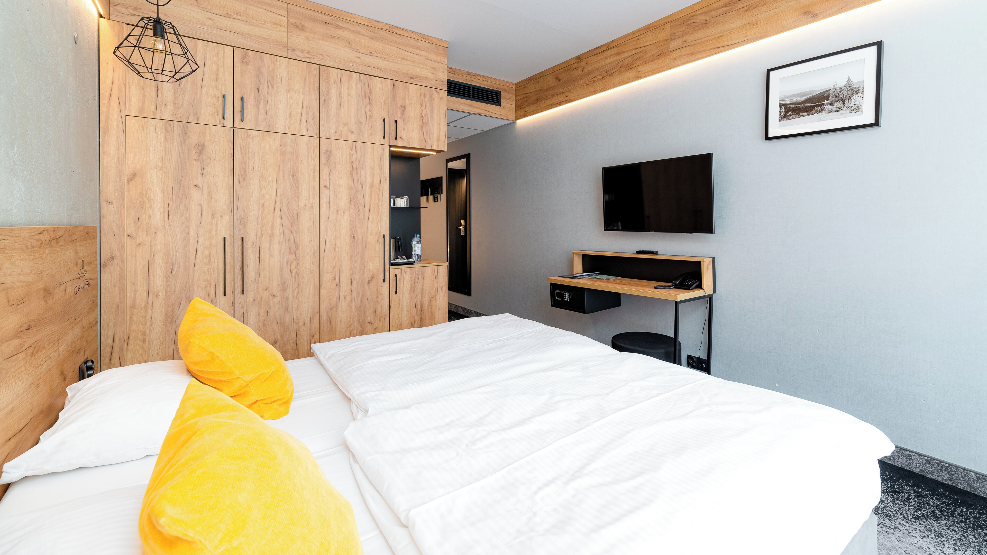 Ein modernes Hotelzimmer mit Bett, Holzschrank, Schreibtisch und einem Wand-Fernseher.