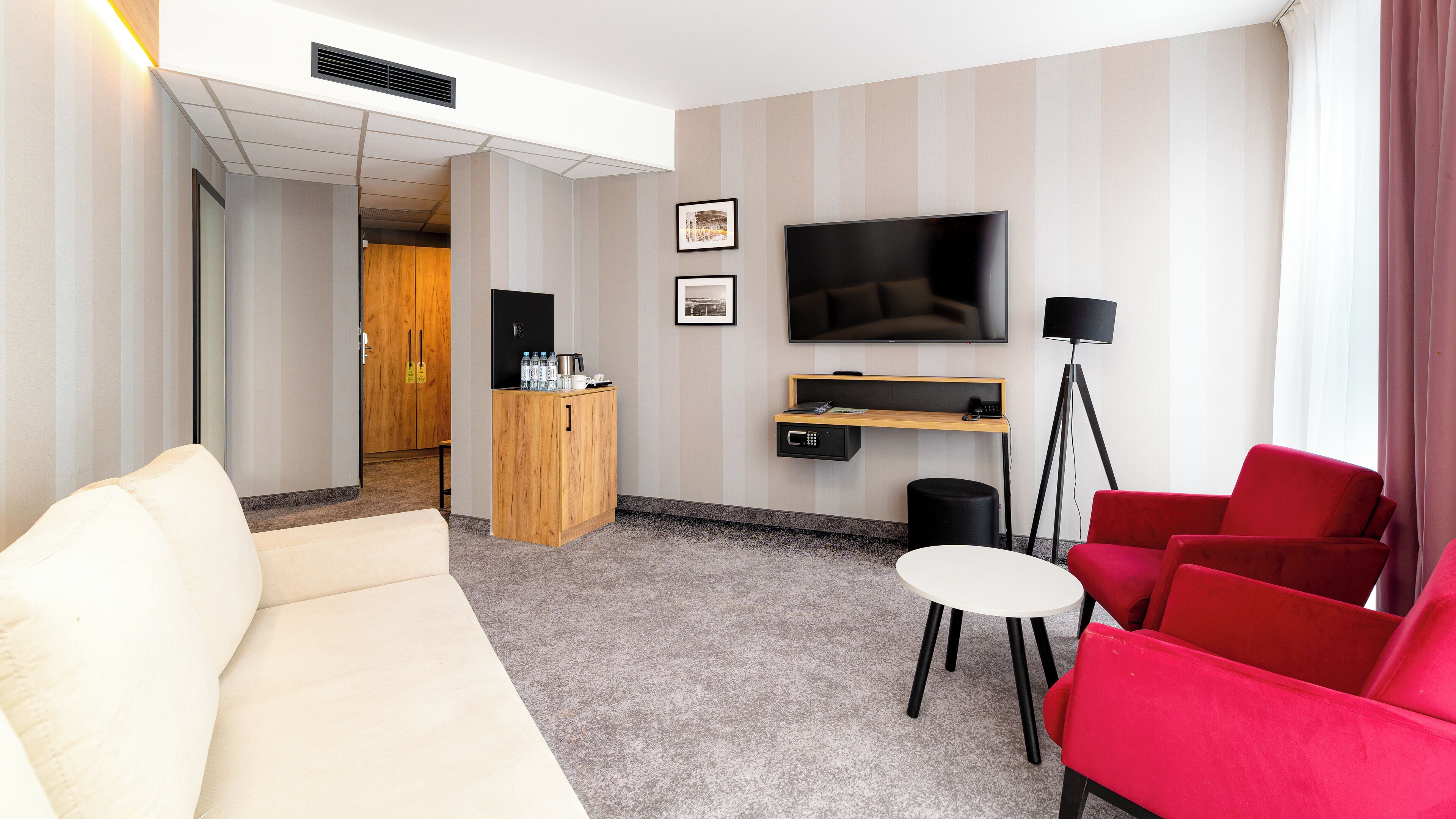 A hotel room with a sofa, red chairs, a TV, a small table, a cabinet, and a floor lamp.