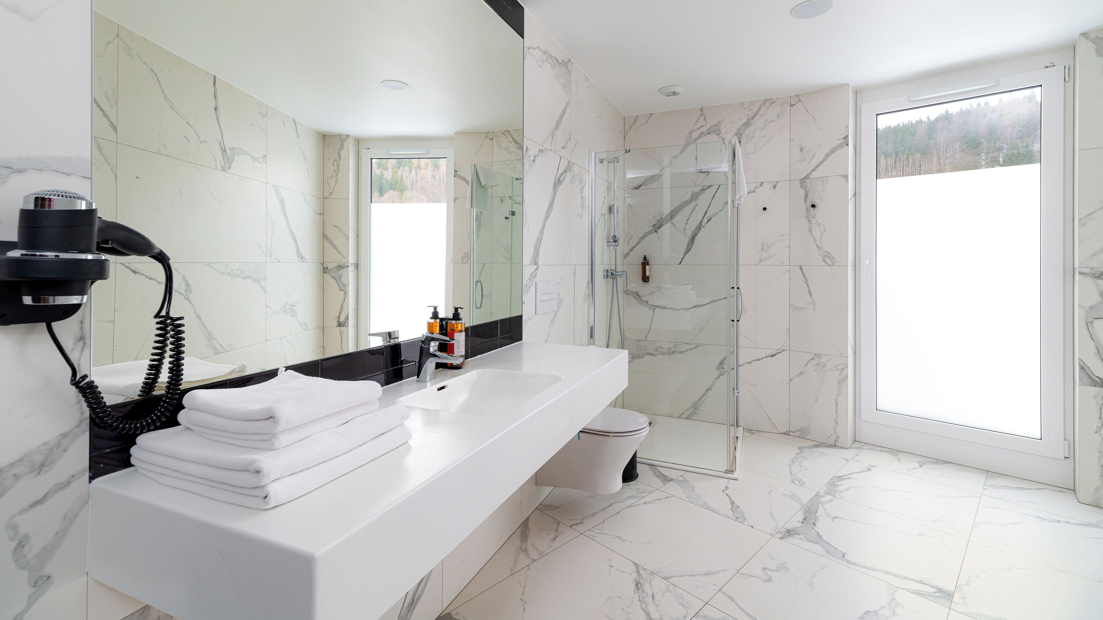 A modern bathroom with a white marble vanity, a glass shower enclosure, and a view of a forest through the window.
