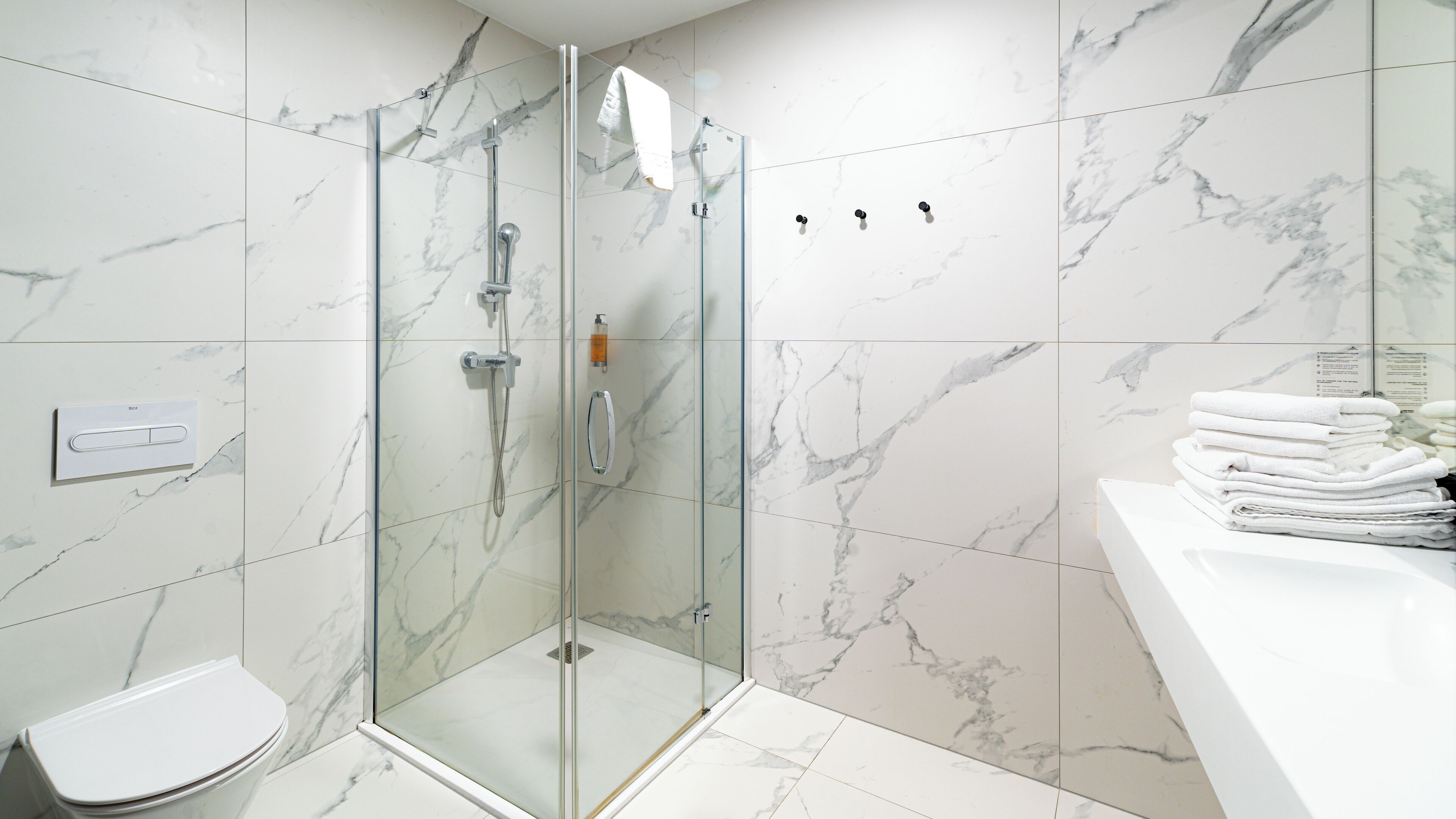 Une salle de bain moderne avec une douche à parois de verre, des carreaux de marbre blanc, un lavabo blanc et une pile de serviettes blanches.