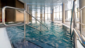 Una piscina interior con agua cristalina, barandillas de metal y sillones de descanso junto a la piscina.