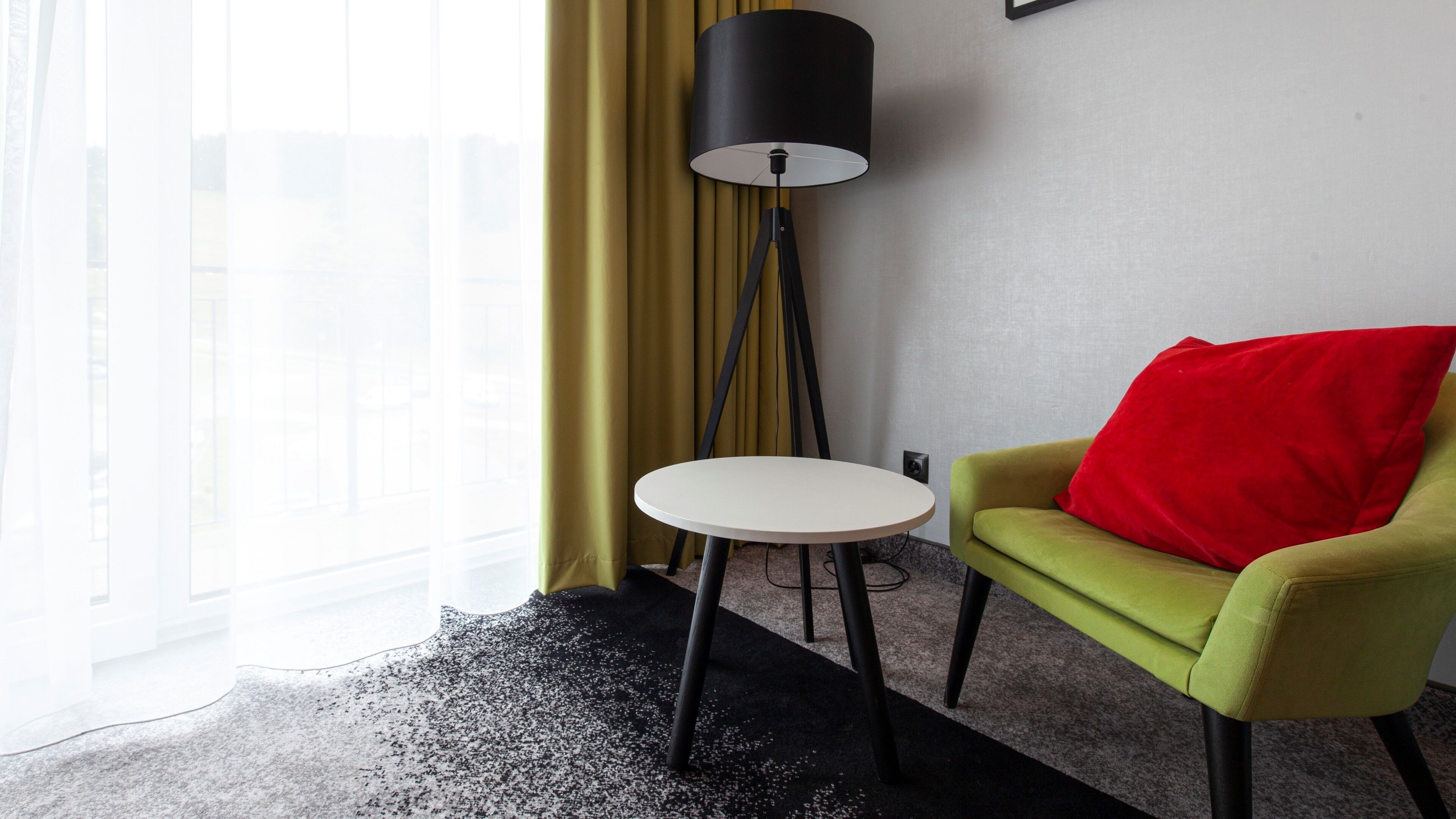 A hotel room with a green armchair, a white round table, a black floor lamp, and a view of a building through large windows.