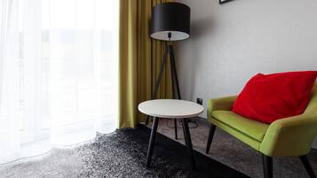 Une chambre d’hôtel avec un fauteuil vert, une table ronde blanche, un lampadaire noir et une vue sur un immeuble par de grandes fenêtres.