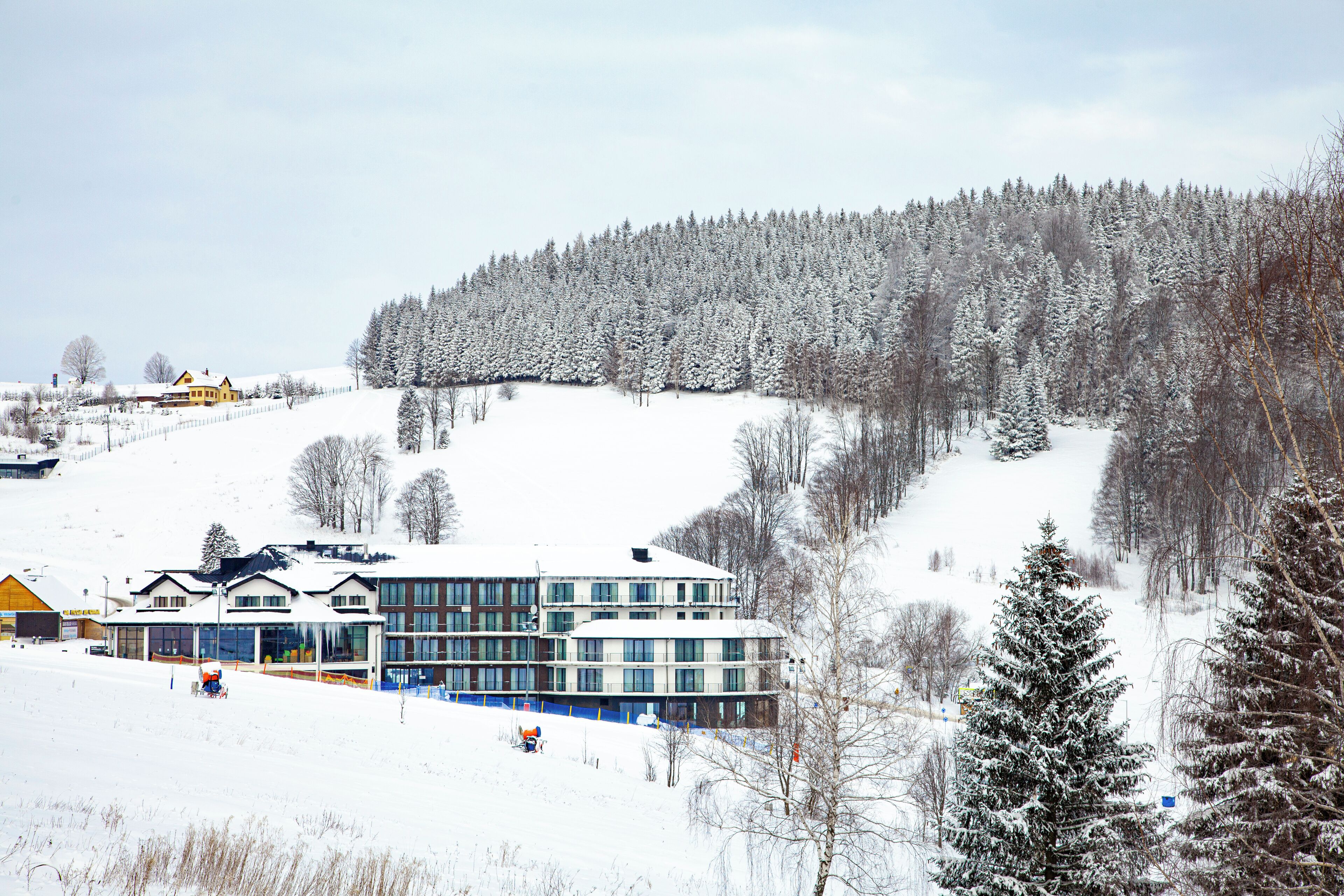 Eine verschneite Landschaft mit einem Gebäude mit mehreren Fenstern und einem flachen Dach, umgeben von schneebehangenen Bäumen und einem klaren Himmel.