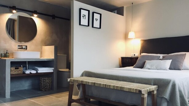 Un dormitorio moderno con cama, mesita de noche, banco y un escritorio pequeño con espejo y espacio de almacenamiento.