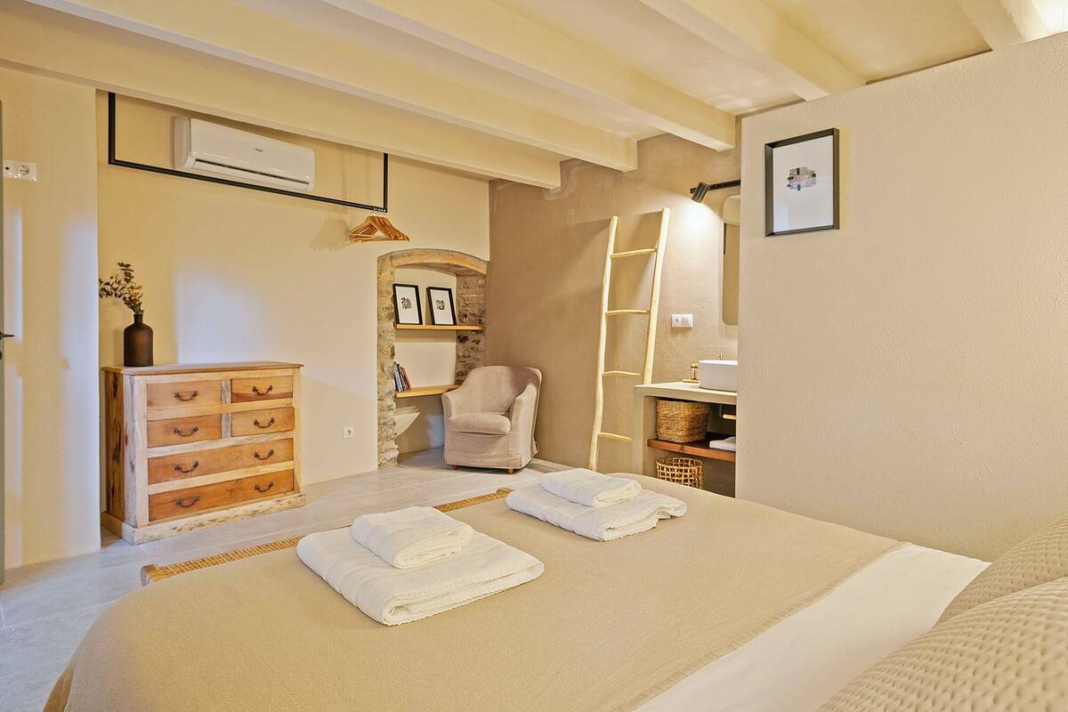 A bedroom with a bed, a wooden dresser, a chair, a shelf with pictures, and an air conditioning unit.