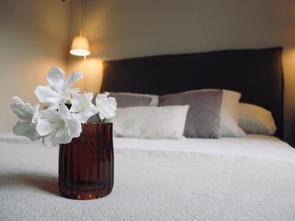 A hotel room with a bed, pillows, a vase with white flowers, and a wall-mounted light.