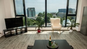 A modern living room with a large window, a flat-screen TV, a white armchair, a small table with a bottle and glasses, and two potted plants. - Diamond View Apartments (Constanta)