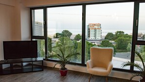 A modern living room with a large window, a flat-screen TV, a comfortable armchair, and a potted plant. - Diamond View Apartments (Constanta)