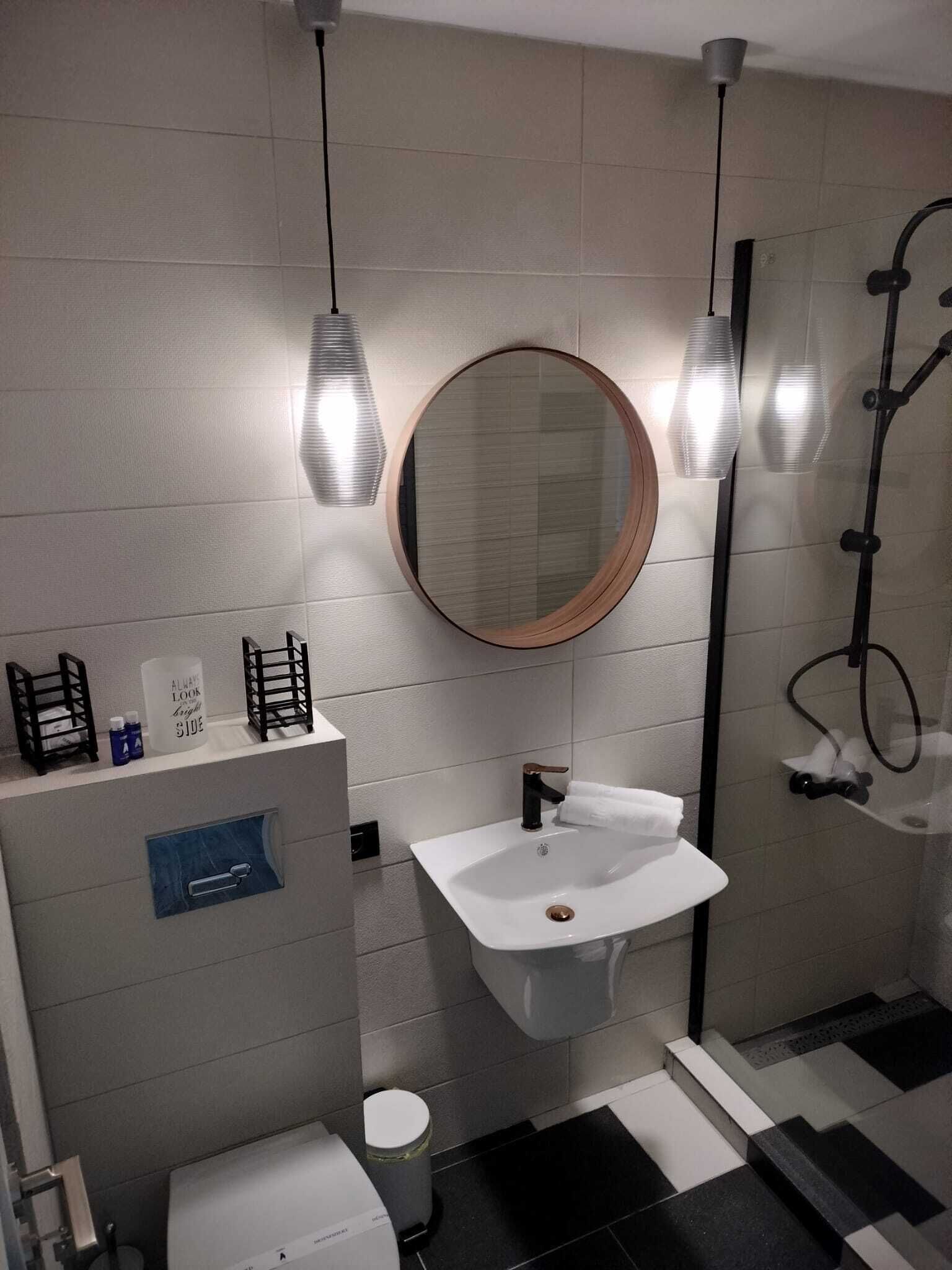 A modern bathroom with a white sink, a round mirror, and a towel rack. There are two black towel racks on the wall and a small trash bin.