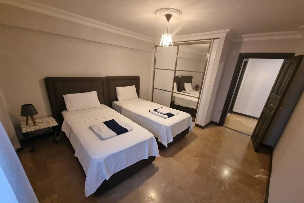 A hotel room with two beds, a nightstand, a lamp, a mirror, and a door.