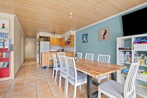 Ein Essbereich mit Holztisch und weißen Stühlen, eine Küche mit Holzausstattung und ein Bücherregal mit Kinderbüchern.