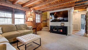 Un salon chaleureux avec un plafond en bois, un canapé beige, un pouf capitonné, un meuble de rangement noir pour appareils électroniques et un escalier menant à un autre niveau.