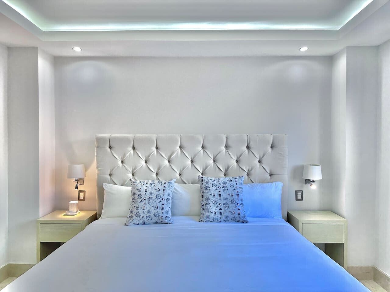 Una cama bien hecha con un cabecero capitonado, dos mesitas de noche con lámparas y almohadas estampadas.