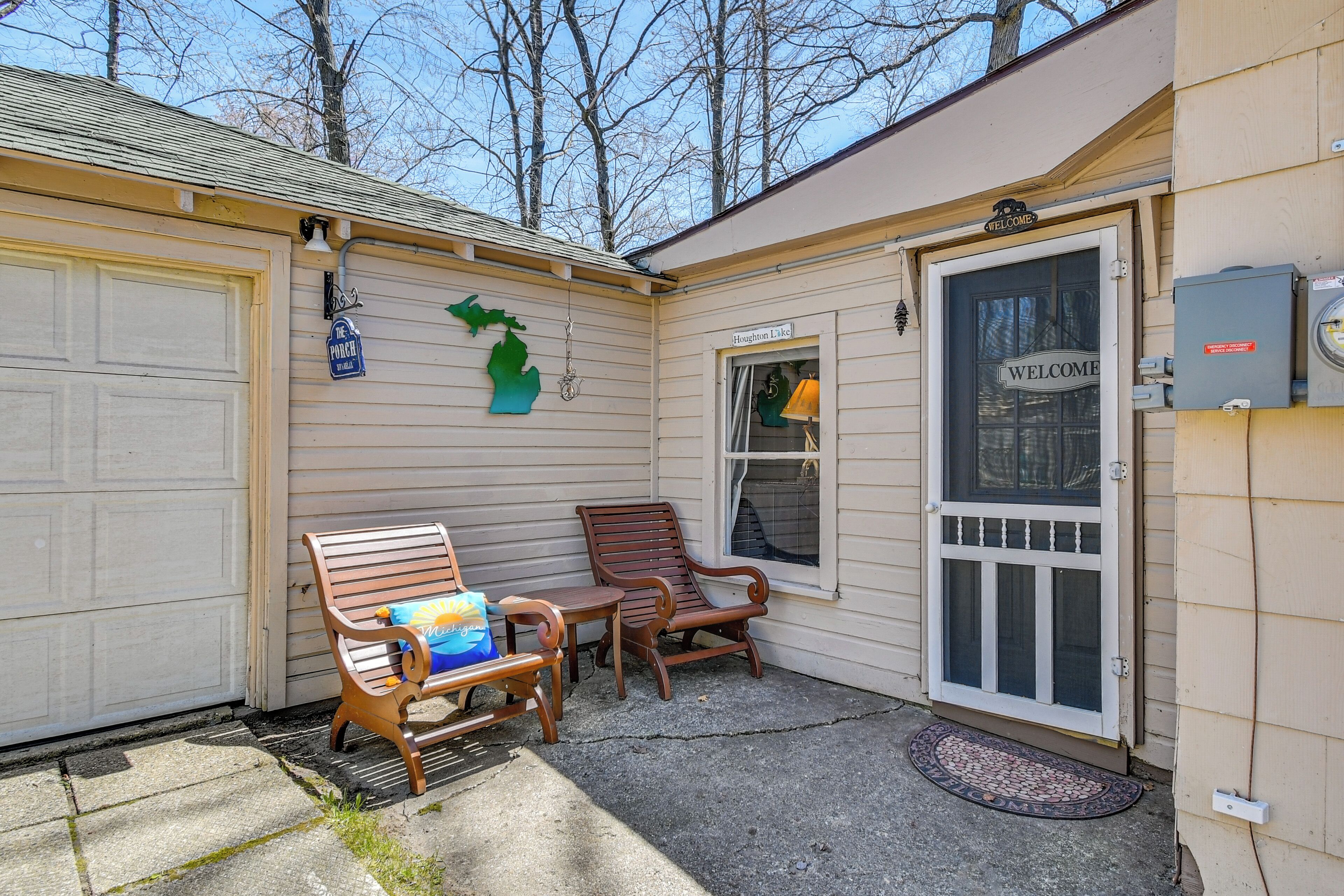 A garage door, a porch with a bench and a chair, a welcome sign, and a window with a view of trees.