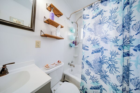 A bathroom with a white sink, a toilet, a bathtub, and a shower curtain with a marine life pattern. There are wooden shelves with toiletries and a mirror above the sink.