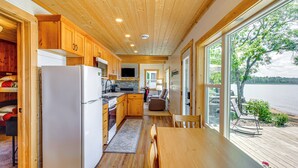 A cabin interior with wooden floors, a kitchen with a refrigerator, stove, and wooden cabinets, a dining area with a table and chairs, and a large window overlooking a lake.