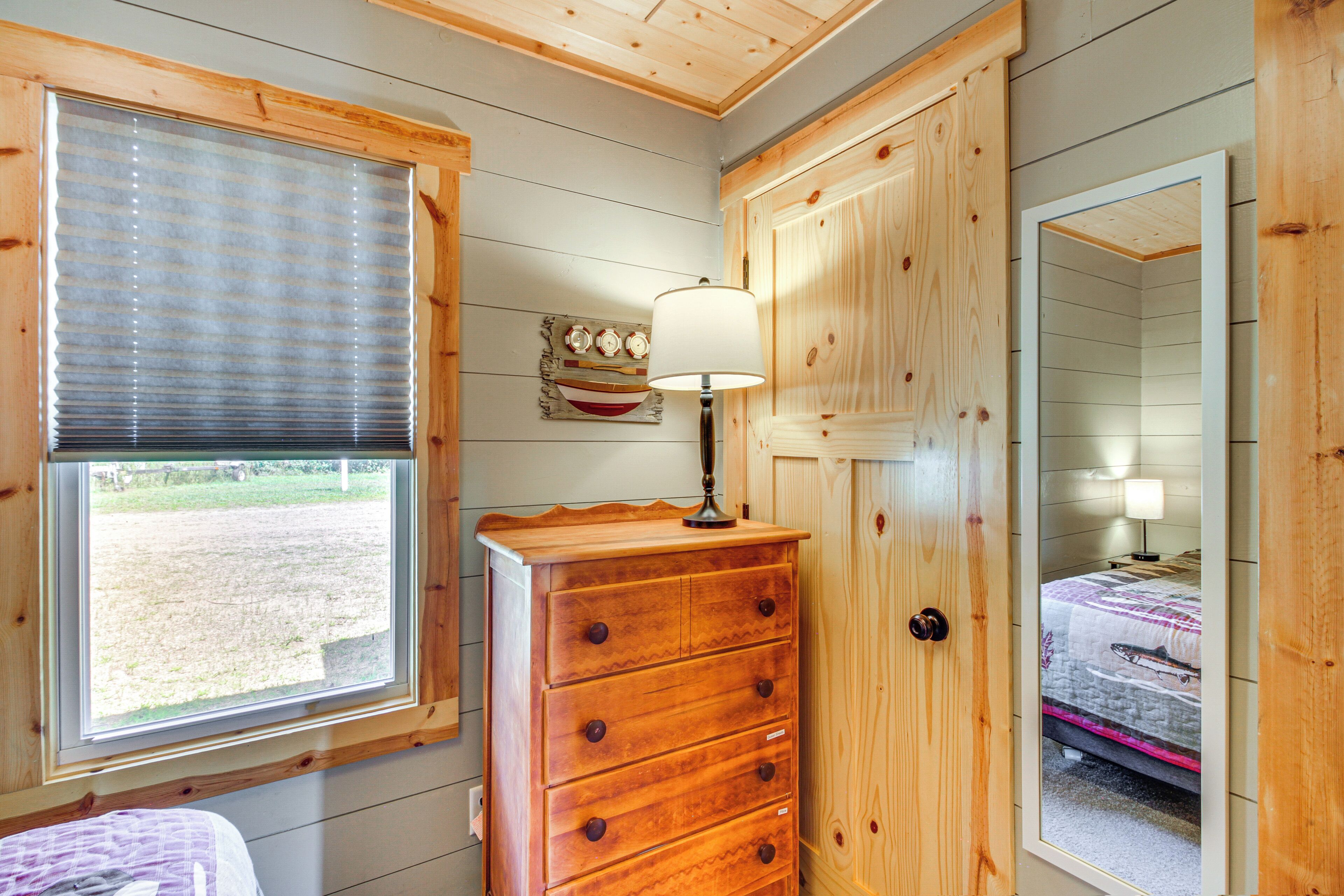 A wooden dresser with drawers, a window with blinds, and a mirror reflecting a bed with a pillow and a lamp on a bedside table.