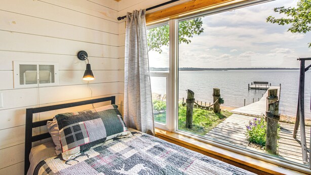 A bedroom with a large window, a bed with a quilt, a wall-mounted lamp, and a view of a dock and water.