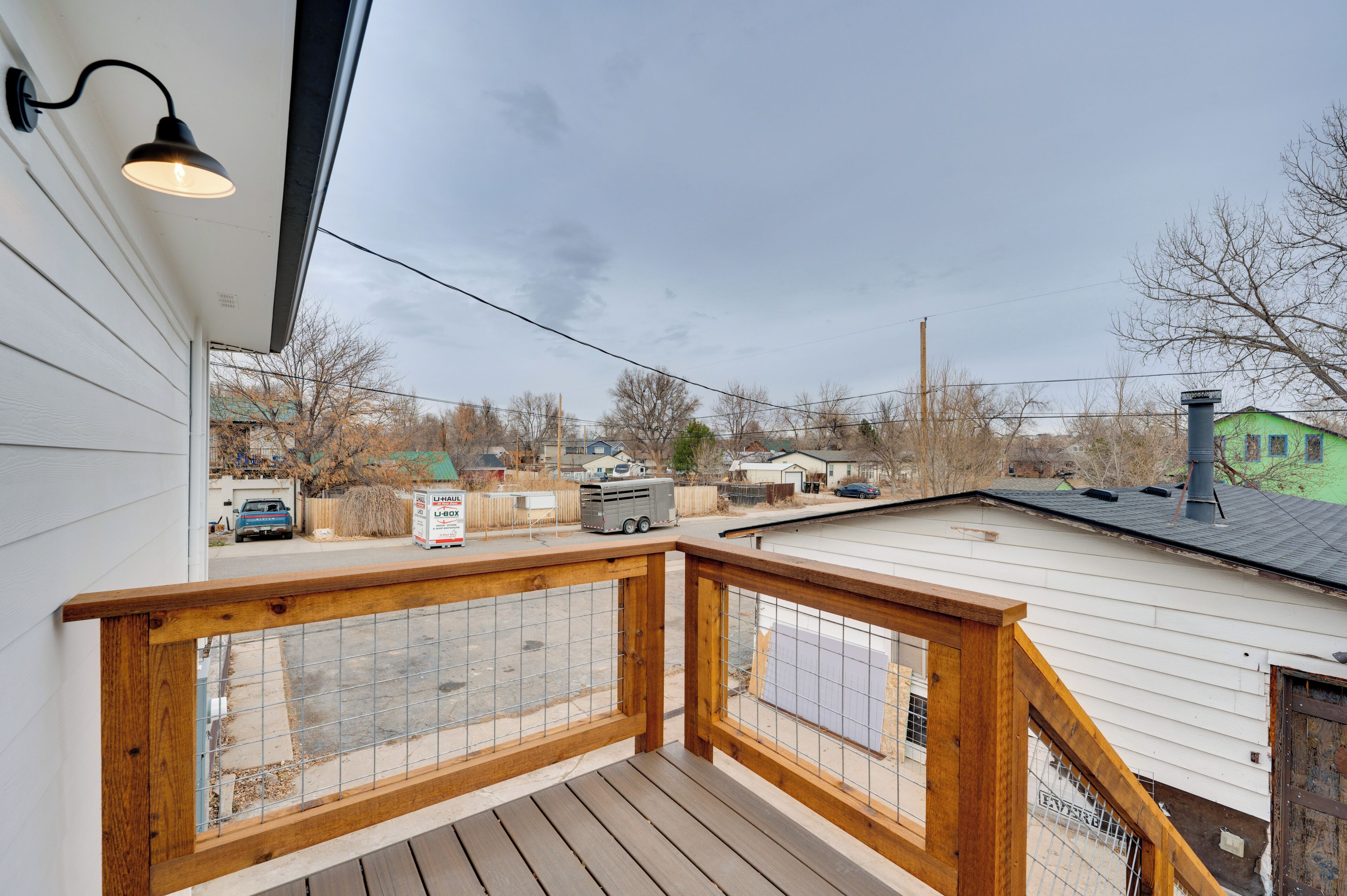 A wooden deck with a metal railing, a hanging light fixture, and a view of a residential area with houses and a parked vehicle.